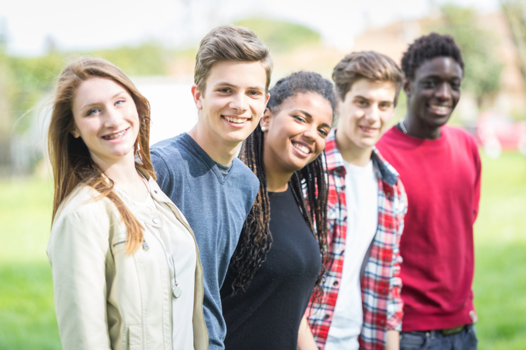 A group of 5 teenagers in a field, standing in a line and smiling at the camera.